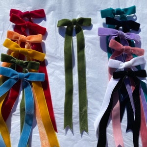 May include: Assortment of velvet ribbon hair bows in various colors, including red, orange, yellow, green, blue, purple, pink, and black. The bows are tied with long tails and are arranged in rows on a white surface.