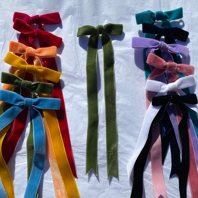 May include: Assortment of velvet ribbon hair bows in various colors, including red, orange, yellow, green, blue, purple, pink, and black. The bows are tied with long tails and are arranged in rows on a white surface.