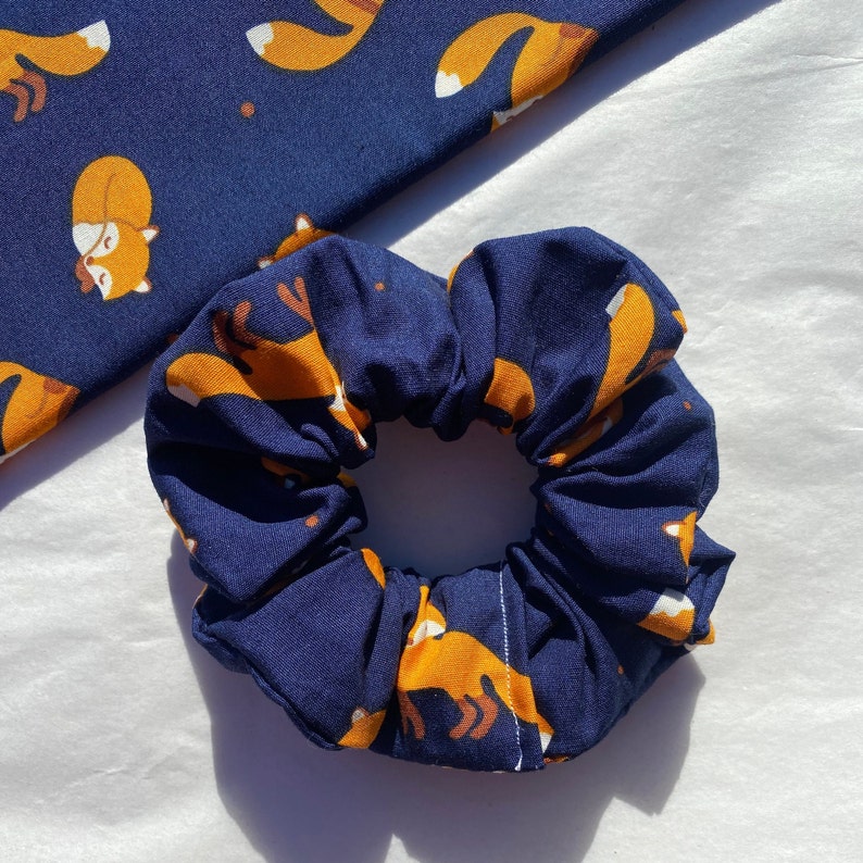 May include: A navy blue scrunchie with a repeating pattern of orange foxes. The foxes are in a sleeping position.