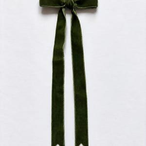 Pode incluir: Um laço de cabelo de veludo verde-oliva escuro com um nó central e duas fitas longas pendentes. O tecido de veludo tem uma aparência macia e texturizada, e as extremidades das fitas são cortadas em um formato clássico de V invertido.