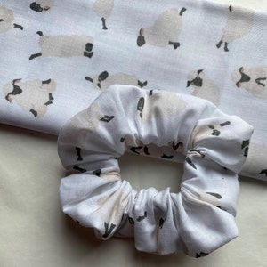 Puede incluir: Scrunchie blanco con un patr&oacute;n repetido de ovejas marrones y blancas. El scrunchie est&aacute; hecho de tela suave y fruncida.