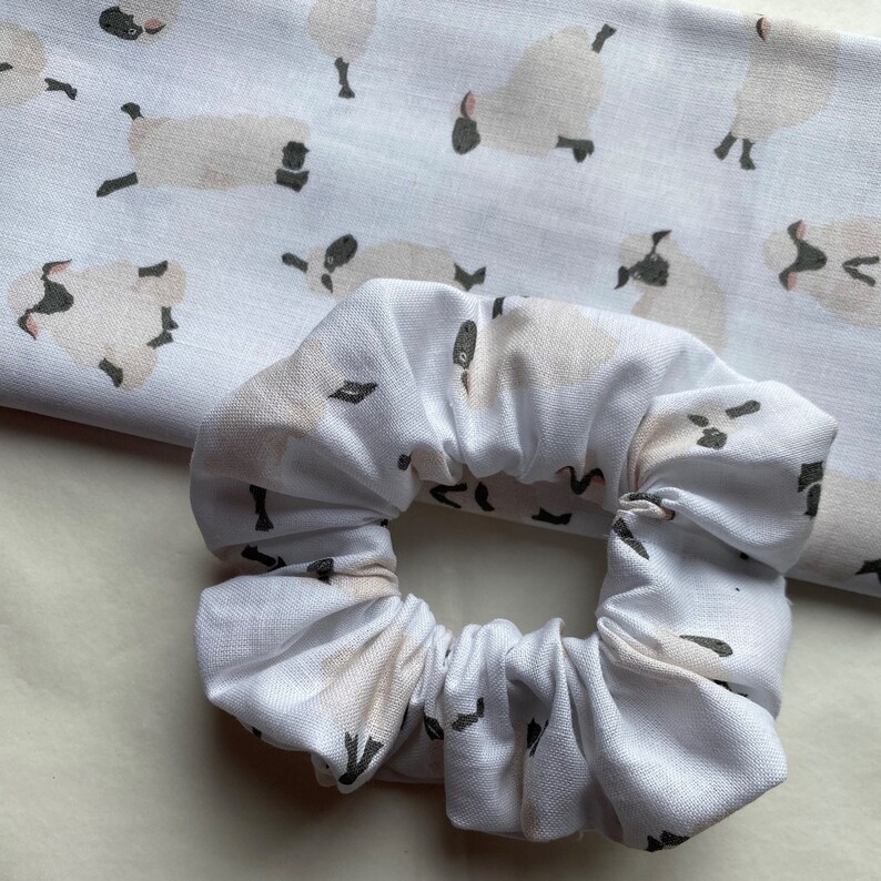 Puede incluir: Scrunchie blanco con un patr&oacute;n repetido de ovejas marrones y blancas. El scrunchie est&aacute; hecho de tela suave y fruncida.