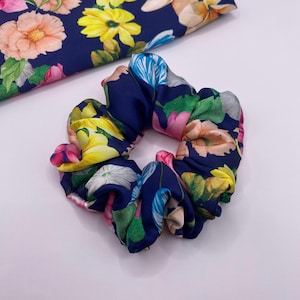 May include: A navy blue scrunchie with a colourful floral design, including yellow, pink, and peach flowers. The scrunchie is made from a soft, silky material. A matching fabric piece is in the background.