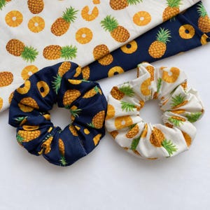 May include: Two fabric hair scrunchies featuring a pineapple pattern. One scrunchie is dark navy blue with yellow and green pineapples, and the other is off-white with a matching pineapple print. Both are displayed on matching fabric pieces.