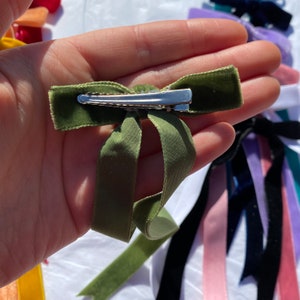May include: A green velvet hair bow with a silver clip. The bow is tied in a simple knot and has long tails.