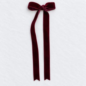 May include: A deep burgundy velvet ribbon tied into a bow with two long, straight tails extending downward. The fabric has a soft, plush texture and a rich, dark red color, featuring clean, finished edges along the entire length of the ribbon.