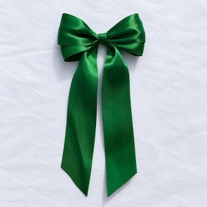 Puede incluir: Un lazo para el cabello de color verde esmeralda vibrante, confeccionado en tela de satén suave y brillante. El accesorio presenta un nudo estructurado en el centro con dos cintas largas y fluidas que caen, mostrando un color verde intenso.