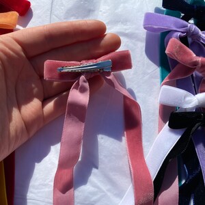 May include: A pink velvet ribbon hair bow with a silver clip. The bow is tied in a simple knot and the ends of the ribbon are long.