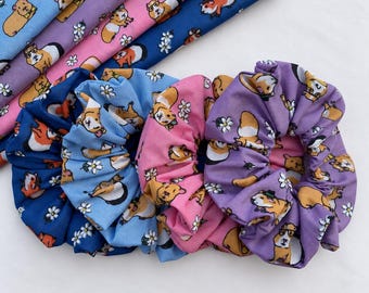 Puede incluir: Tres coleteros de tela en azul, rosa y morado, cada uno con un patrón repetitivo de cobayas de dibujos animados y pequeñas margaritas blancas. Los coleteros están dispuestos en fila, con tiras de tela plegadas a juego detrás de ellos.