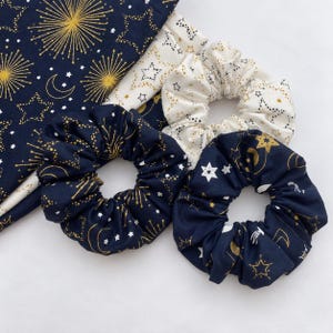 May include: Two dark navy blue fabric hair scrunchies with gold star, moon, and firework patterns, one cream-colored fabric hair scrunchie with gold and black star patterns, and a matching piece of dark navy blue fabric with gold firework designs.