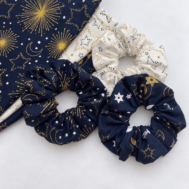May include: Two dark navy blue fabric hair scrunchies with gold star, moon, and firework patterns, one cream-colored fabric hair scrunchie with gold and black star patterns, and a matching piece of dark navy blue fabric with gold firework designs.