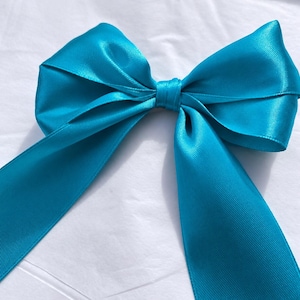 Op de afbeelding: Een fel turquoise satijnen lint gestrikt tot een decoratieve strik. De strik heeft een centrale knoop met twee symmetrische lussen en twee lange, neerhangende uiteinden, wat zorgt voor een gladde, reflecterende textuur en een levendige kleur.