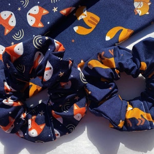 May include: Two navy blue scrunchies with a fox and star pattern. The scrunchies are made of soft fabric and are perfect for holding hair in place.