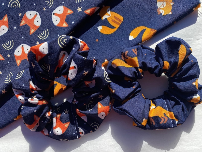 May include: Two navy blue scrunchies with a fox and star pattern. The scrunchies are made of soft fabric and are perfect for holding hair in place.