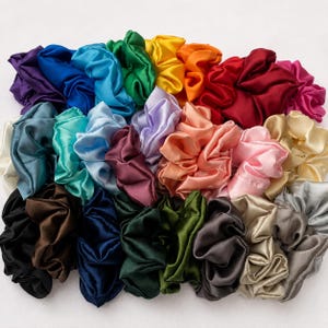 May include: A collection of numerous satin hair scrunchies in a wide variety of colours including purple, blue, green, yellow, orange, red, pink, brown, black, grey, and cream. Each scrunchie features a gathered, pleated fabric design.