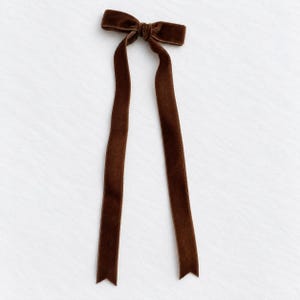 May include: A dark brown velvet hair bow featuring a central knot and two long, straight ribbon tails that extend downwards. The fabric has a soft, textured appearance with finished edges, creating a simple and classic accessory design.