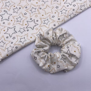 Puede incluir: Un scrunchie blanco con un patr&oacute;n de estrellas doradas y negras. El scrunchie est&aacute; hecho de un tejido suave y el&aacute;stico, perfecto para sujetar el cabello.