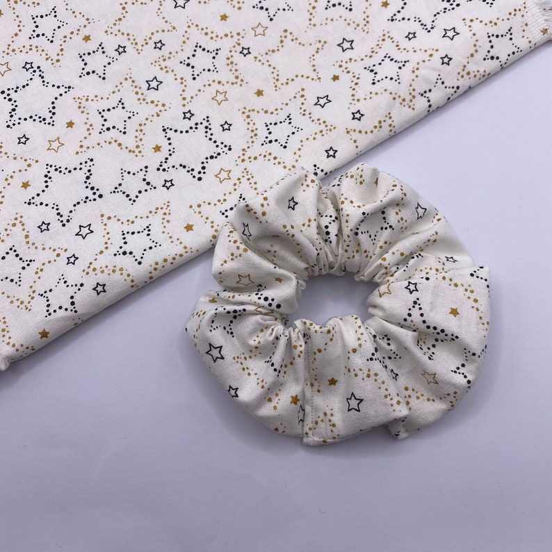 Puede incluir: Un scrunchie blanco con un patr&oacute;n de estrellas doradas y negras. El scrunchie est&aacute; hecho de un tejido suave y el&aacute;stico, perfecto para sujetar el cabello.
