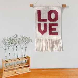 May include: A cream-coloured macrame wall hanging with the word "LOVE" in burgundy. It has a wooden dowel and fringe. A wooden test tube holder with flowers is on a wooden surface. The wall is white.