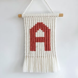 May include: A macrame wall hanging featuring the letter "A" in a terracotta color against a cream background. The piece is suspended from a wooden dowel with a cream-colored cord and has a fringe at the bottom.