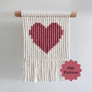 May include: A macramé wall hanging with a heart design in red and white. The heart is central on a rectangular panel with a fringed bottom edge. It hangs from a wooden dowel. The image also includes a PDF Pattern graphic.
