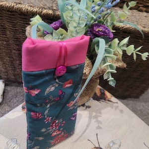 May include: A teal blue fabric pouch with a pink flap and a floral print. The pouch has a pink zipper and a pink button. The pouch is sitting on a surface with a bee pattern.