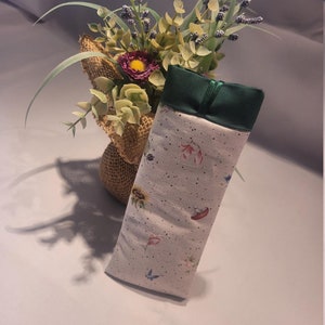 May include: A rectangular, soft fabric case with a floral pattern featuring sunflowers, butterflies, and other small flowers. The top is a solid dark green, and the case is slightly angled. A bouquet of flowers in a burlap wrap is in the background.