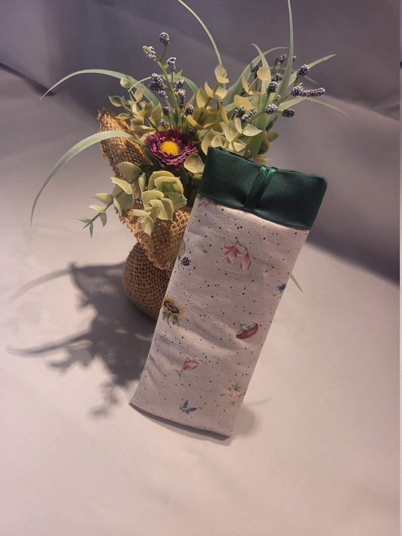 May include: A rectangular, soft fabric case with a floral pattern featuring sunflowers, butterflies, and other small flowers. The top is a solid dark green, and the case is slightly angled. A bouquet of flowers in a burlap wrap is in the background.