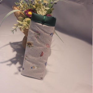 May include: A rectangular, fabric glasses case with a floral and insect pattern. The case is light gray with a green top and a green ribbon. The pattern includes sunflowers, mushrooms, and butterflies. The case is displayed next to a burlap-wrapped vase with flowers.
