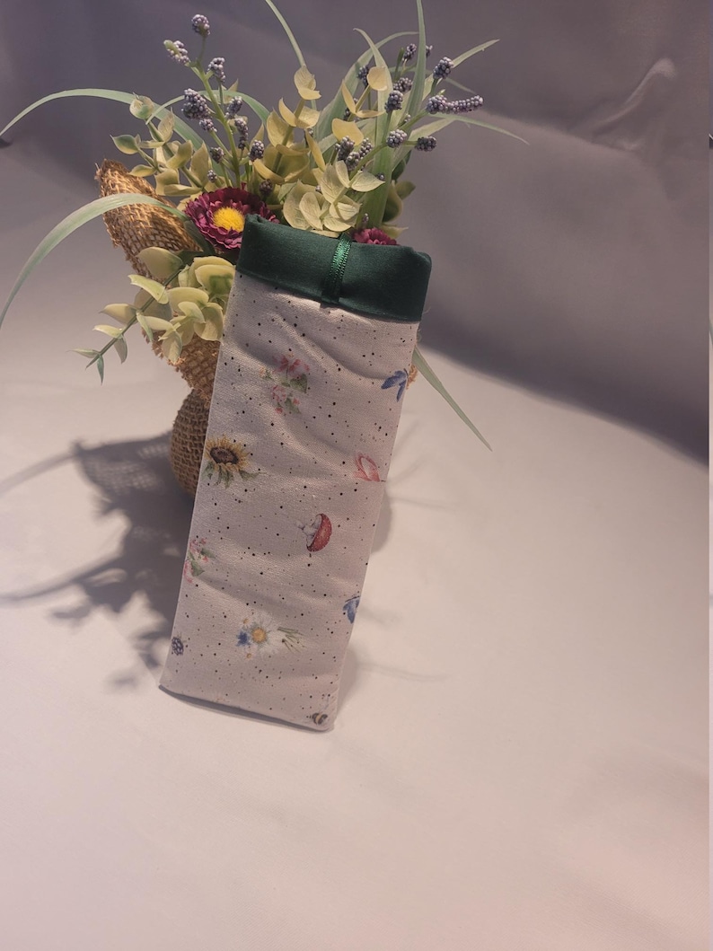 May include: A rectangular, fabric glasses case with a floral and insect pattern. The case is light gray with a green top and a green ribbon. The pattern includes sunflowers, mushrooms, and butterflies. The case is displayed next to a burlap-wrapped vase with flowers.