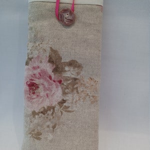 May include: A rectangular eyeglasses case with a floral rose pattern in pink and brown on a beige linen fabric. The top is a solid off-white fabric. A pink ribbon and a decorative button secure the case.