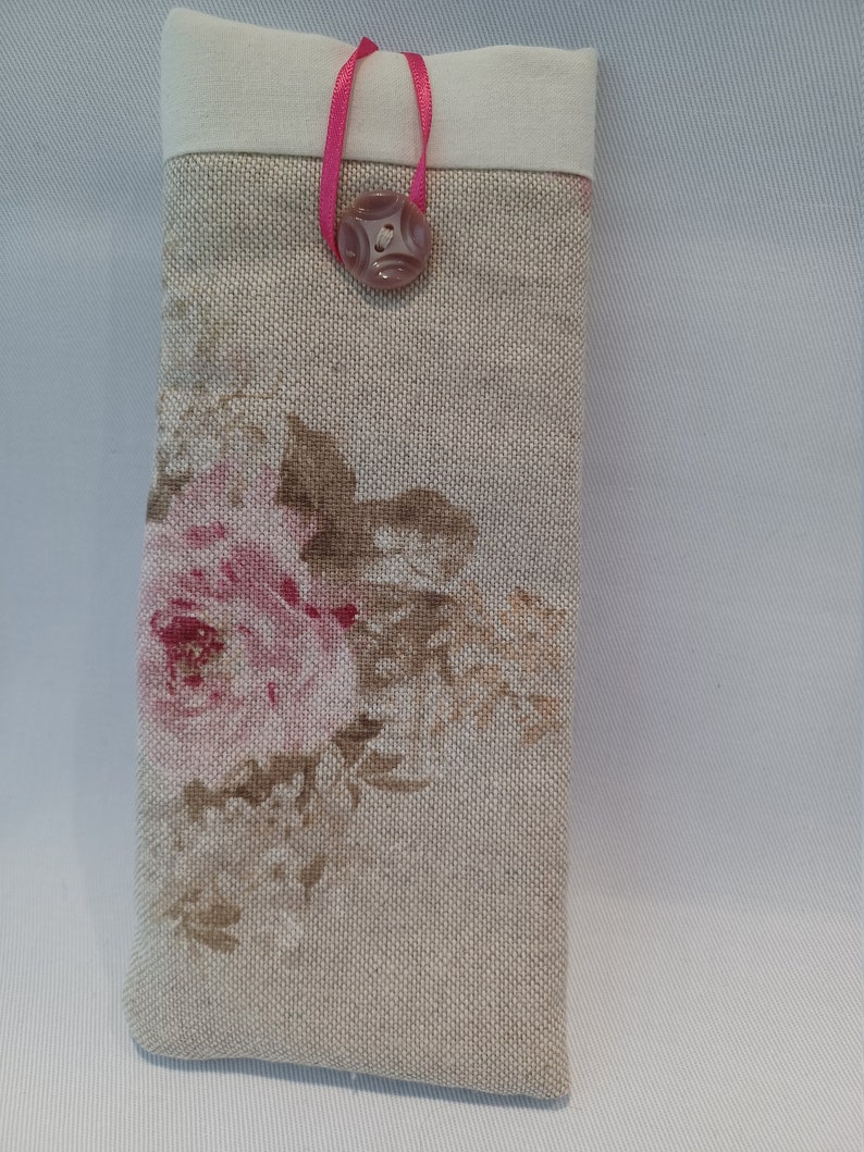 May include: A rectangular eyeglasses case with a floral rose pattern in pink and brown on a beige linen fabric. The top is a solid off-white fabric. A pink ribbon and a decorative button secure the case.