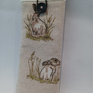 May include: A beige fabric glasses case featuring a repeating pattern of brown hares in a grassy field. The case has a cream-colored top and is secured with a black cord and a star-shaped button.