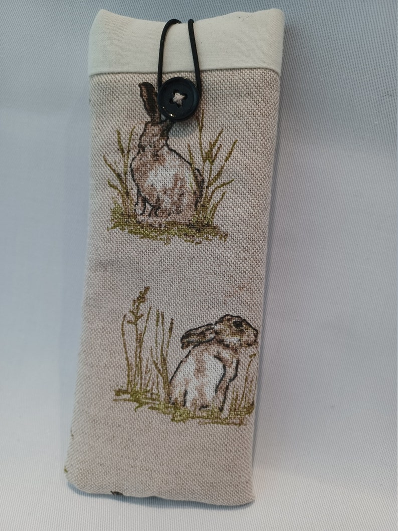 May include: A beige fabric glasses case featuring a repeating pattern of brown hares in a grassy field. The case has a cream-colored top and is secured with a black cord and a star-shaped button.