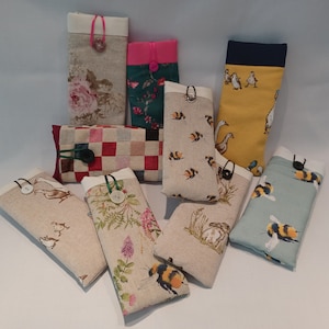 May include: A collection of fabric cases in various designs. Some cases feature floral patterns, bees, geese, and a checkered pattern. Each case has a contrasting top and a button closure. The cases are designed to hold glasses or other small items.