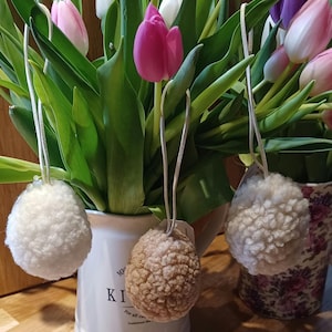 May include: A display of pink, white, and purple tulips in white and floral patterned vases. Three fluffy, round, off-white and tan pom-pom ornaments hang in front of the flowers, adding a decorative touch to the spring floral arrangement.