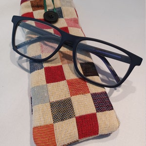 May include: Rectangular navy blue eyeglasses rest on a colorful, checkered glasses case. The case is made of woven fabric with a button closure and a green loop. The glasses have a rectangular frame.