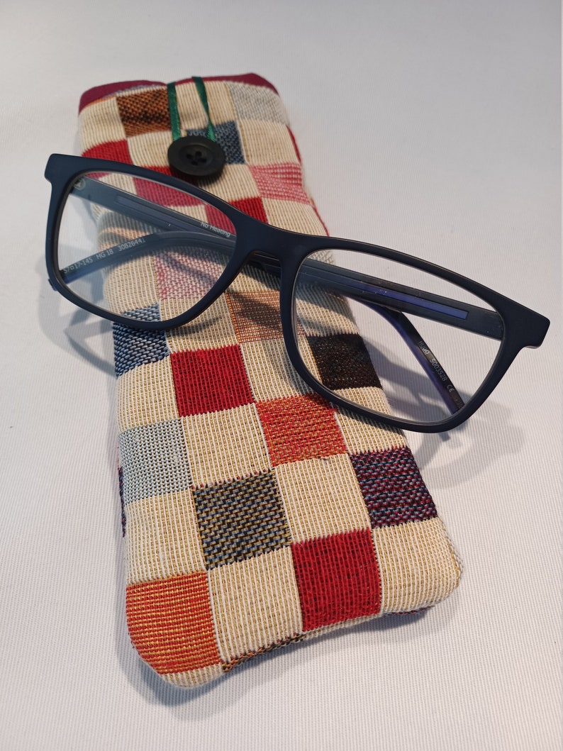 May include: Rectangular navy blue eyeglasses rest on a colorful, checkered glasses case. The case is made of woven fabric with a button closure and a green loop. The glasses have a rectangular frame.