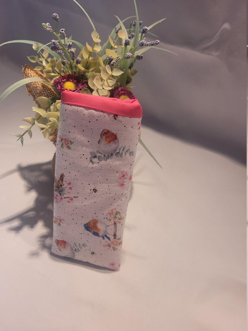 May include: A fabric glasses case with a pink trim and a bird and floral pattern. The case is white with small black dots and features illustrations of birds, flowers, and butterflies. The case is folded and stands upright.
