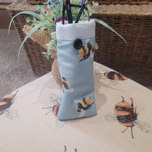 May include: A light blue glasses case with a bumblebee pattern. The case has a white top and a black button closure. Black glasses are inside the case. The case is sitting on a beige surface with a bumblebee pattern.