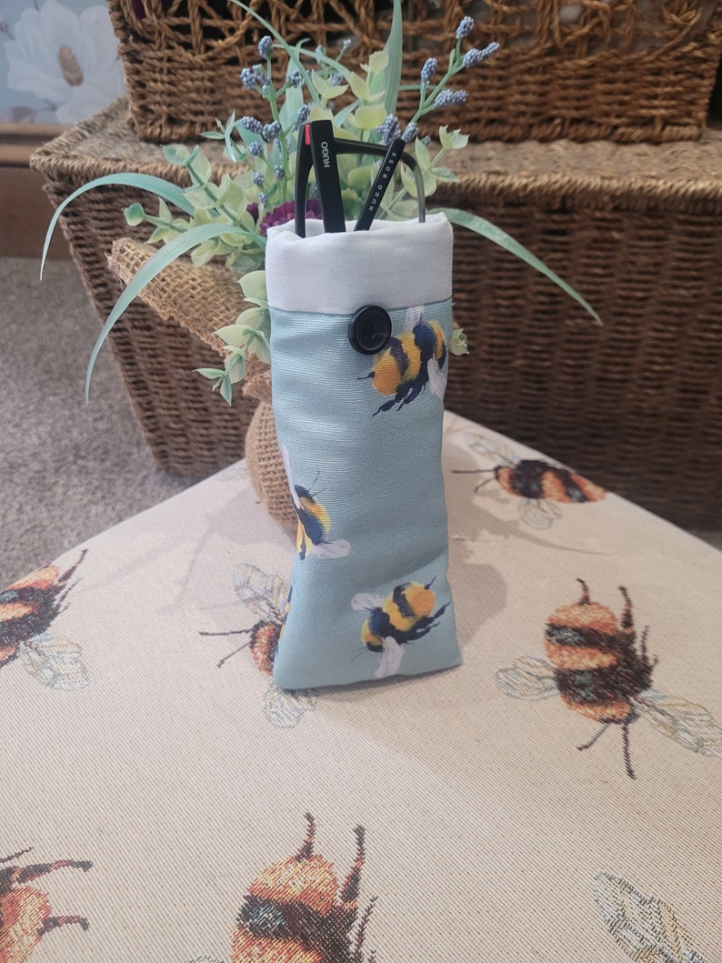 May include: A light blue glasses case with a bumblebee pattern. The case has a white top and a black button closure. Black glasses are inside the case. The case is sitting on a beige surface with a bumblebee pattern.