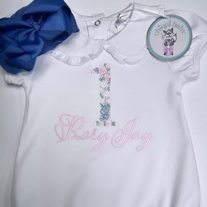 May include: White baby romper with a floral embroidered "1" and the name "Rory Joy" in pink script. A large blue bow is placed near the romper. The romper has a Peter Pan collar and the brand tag "chiqui baby".
