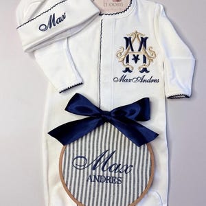 Puede incluir: Conjunto de ropa de bebé blanco con detalles azul marino. Incluye un gorro, un body con iniciales y nombre bordados, y un aro a rayas con el nombre "Max Andres". Un lazo azul marino adorna el conjunto.