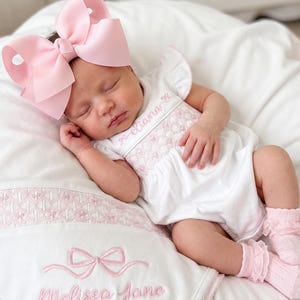 May include: A newborn wearing a white short-sleeved romper with pink smocking and the name Eliana embroidered on the chest, paired with pink ruffled socks and a large light pink hair bow. A matching white blanket features pink embroidery of a bow and the name Melissa Jane.