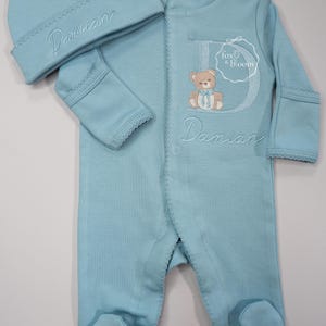 May include: A light blue baby outfit set, including a beanie and a footed onesie. The onesie features a teddy bear applique and the name "Damian" embroidered. The set is from "fox & bloom".