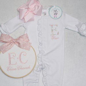 May include: White baby onesie with pink floral embroidery of the letter E and the name "Elissa". A pink bow is on the left side of the image and a white embroidery hoop with the letters "E.C" and the name "Elissa Chaouk" is on the left side of the image. The onesie has ruffles around the neckline and sleeves.