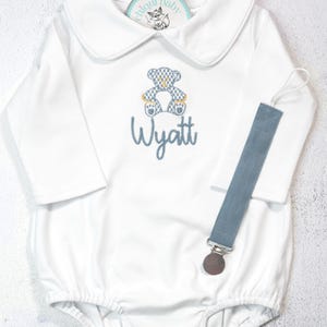 Könnte beinhalten: Weißer Baby-Strampler mit Bubikragen, langen Ärmeln und einem blauen Teddybär-Applikation. Der Name "Wyatt" ist darunter gestickt. Ein blauer Schnullerclip ist befestigt. Der Strampler ist für Babys.