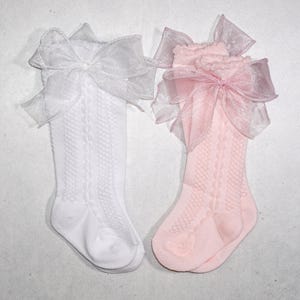 May include: Two pairs of knee-high socks, one white and one pink, each adorned with a large, sheer bow at the top. The socks have a textured, patterned design. The bows are made of a delicate, translucent material.
