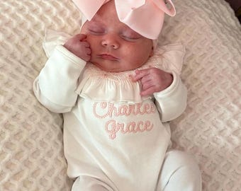 May include: A newborn baby wearing a white onesie with the embroidered name "Charlee Grace" in pink thread. The baby is also wearing a large pink bow headband and is lying on a white textured blanket.
