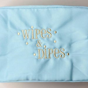 Puede incluir: Una bolsa rectangular azul claro con las palabras "wipes & dipes" bordadas en hilo dorado. La bolsa tiene cierre de cremallera y está destinada a artículos para el cuidado del bebé.